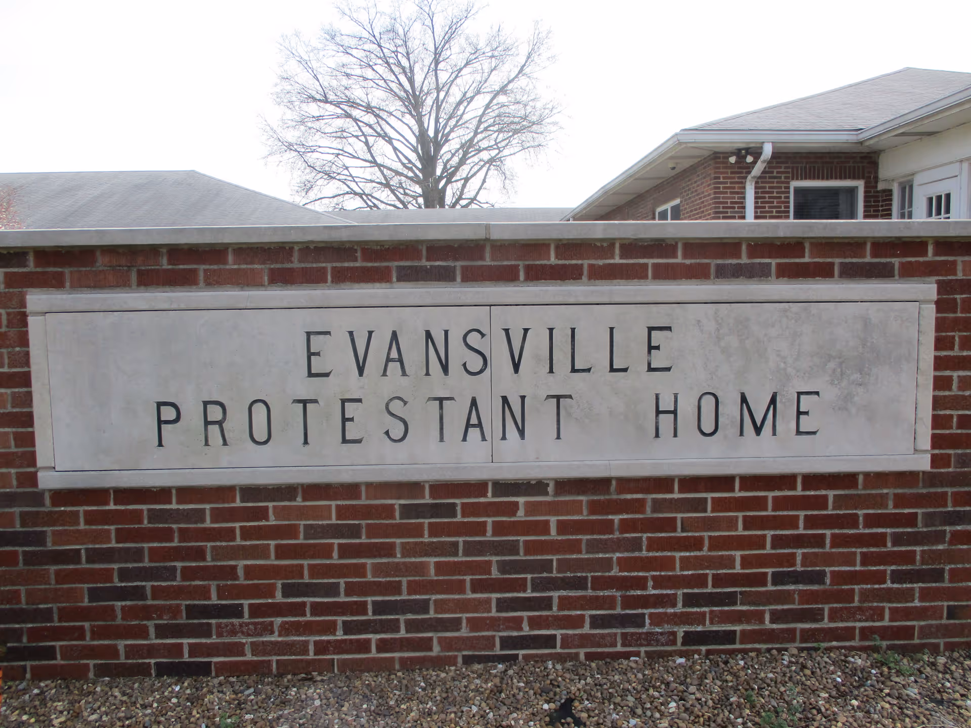 A brick wall with a large rectangular stone plaque embedded in it. The plaque has the text 'EVANSVILLE PROTESTANT HOME' engraved in capital letters. Behind the wall, parts of a building and a leafless tree are visible.