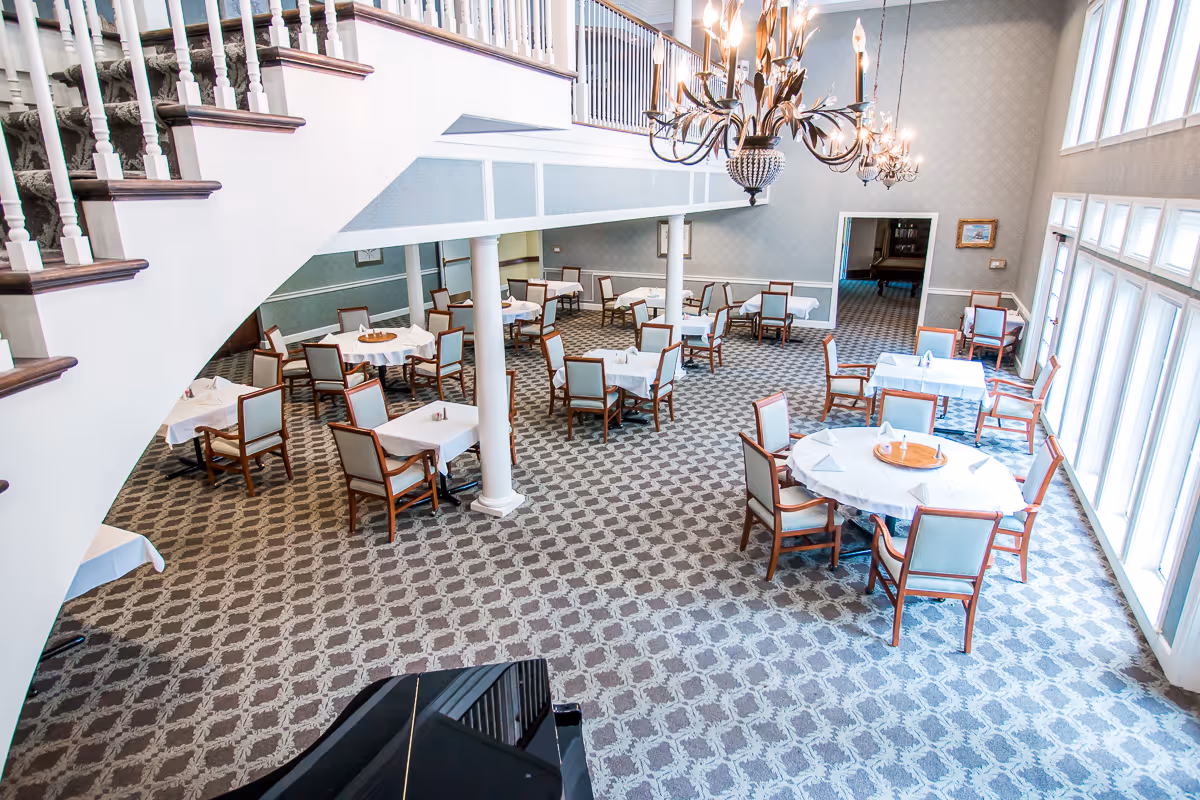 A spacious dining room with multiple round and square tables covered with white tablecloths, surrounded by wooden chairs with light upholstery. The room features a patterned carpet, large windows allowing natural light, a grand chandelier, and a staircase with white railings.