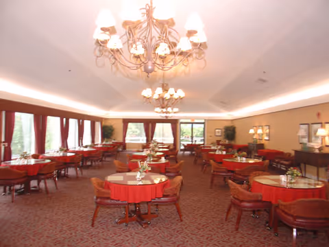 Spacious dining room with multiple round tables covered with red tablecloths, each surrounded by wooden chairs with red cushions. The room features large windows with red curtains allowing natural light, two ornate chandeliers hanging from the ceiling, and decorative plants along the walls.
