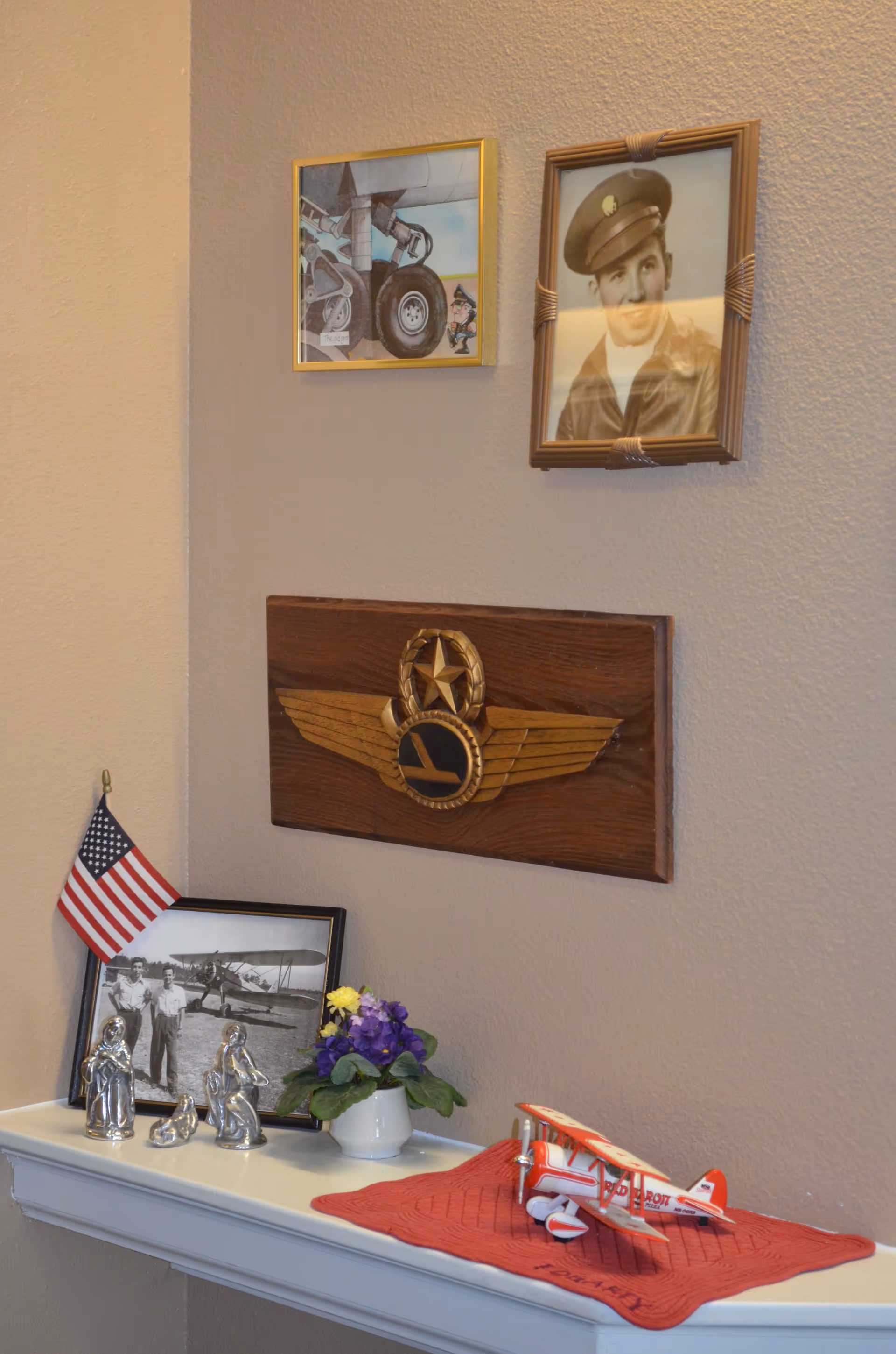 Interior wall display with aviation-themed plaques and framed photos above a shelf holding a model biplane, a small American flag, figurines and a potted plant.