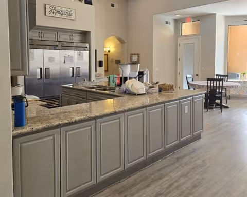 Communal kitchen area with a long granite-topped island, stainless refrigerators, and a small dining table visible in the background.