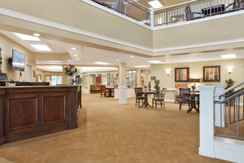 Interior view of a senior living facility lobby area with a wooden reception desk on the left, small round tables with chairs, decorative plants, framed artwork on the walls, and a staircase railing on the right. The space has beige walls, carpeted floors, and a high ceiling with a second-floor balcony visible.