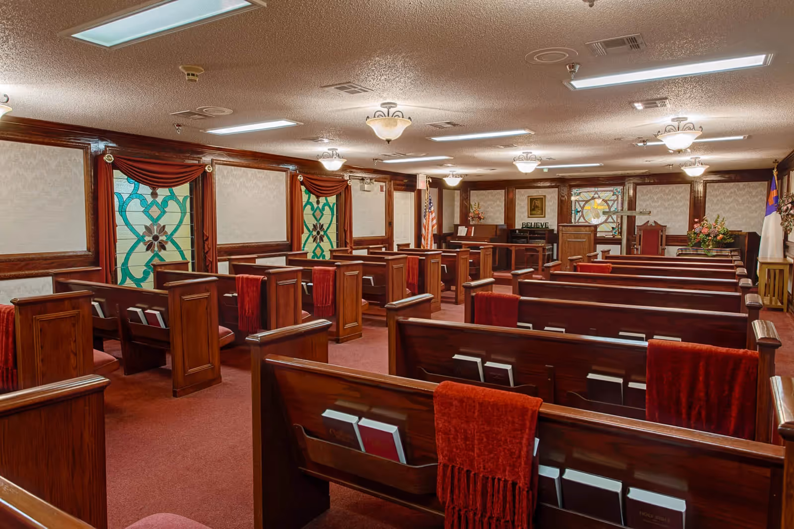 Interior chapel-like room with wooden pews, red carpeting, stained glass windows and an altar at the front.