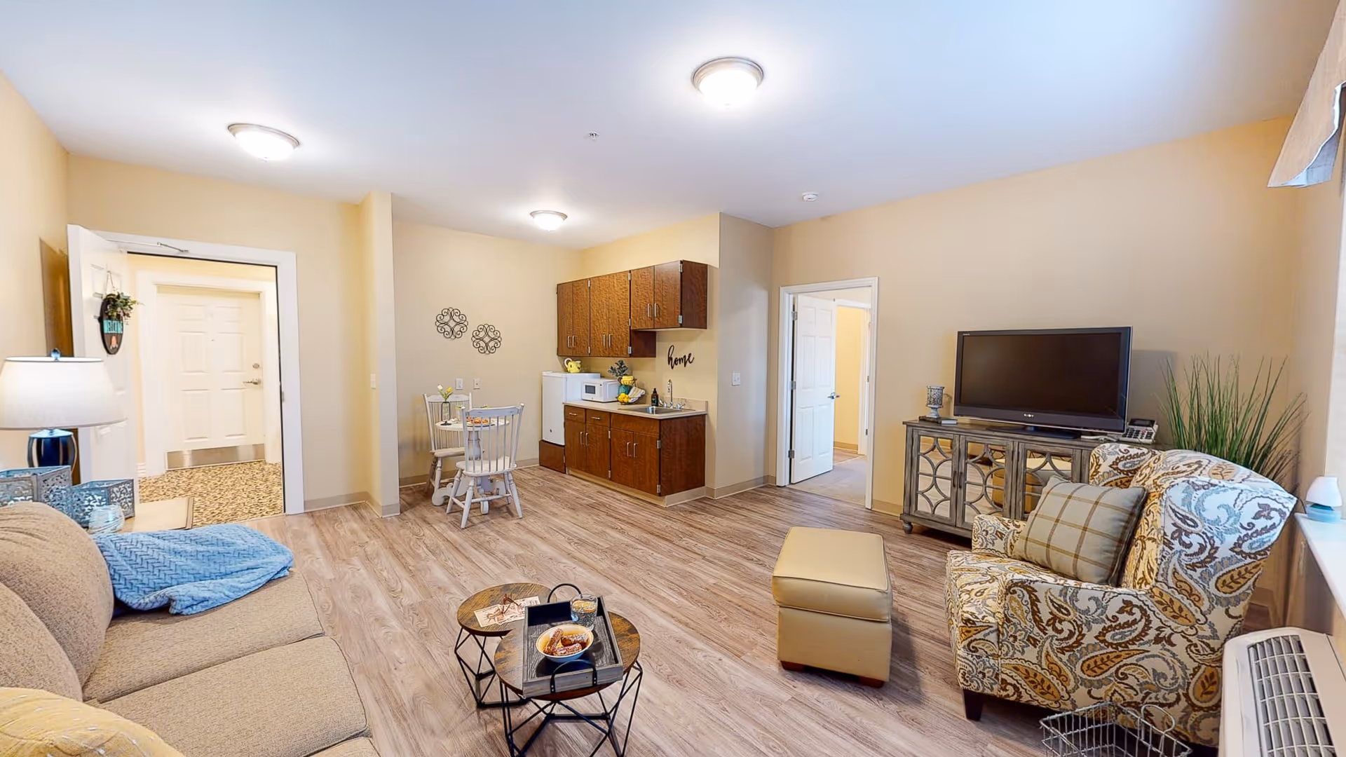A cozy living room area in a senior living facility featuring a beige sofa with a blue throw blanket, a patterned armchair with a plaid pillow, a small ottoman, and a TV on a decorative wooden stand. The room has light wood flooring, beige walls, and a small kitchenette with cabinets, a microwave, and a mini fridge. A small round dining table with two chairs is positioned near the kitchenette. There are two doorways visible, one leading to a hallway and another to a different room.