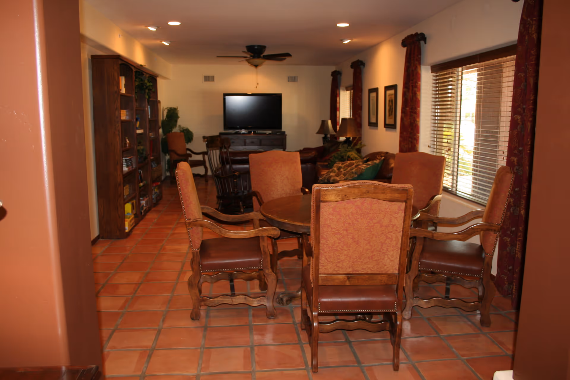 Interior view of a living room in Doubletree Ranchos Assisted Living featuring a round wooden table with five upholstered chairs in the foreground, a tiled floor, a ceiling fan, a flat-screen TV on a wooden cabinet, bookshelves along the left wall, and windows with blinds and red curtains on the right.