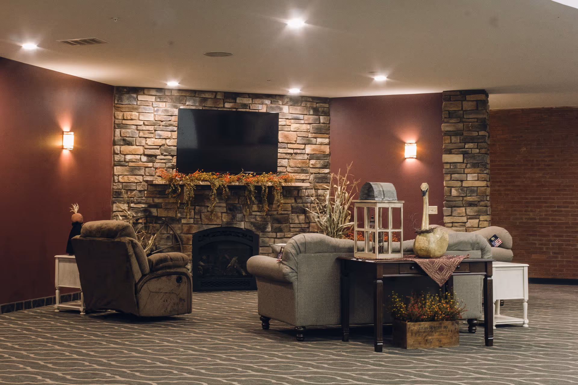 A cozy living room area with a stone fireplace and a mounted flat-screen TV above it. The room features a comfortable gray sofa, a brown recliner chair, a wooden side table with decorative items including a lantern and a pumpkin, and warm wall sconces lighting the space. The walls are painted a deep red color and the floor is carpeted with a patterned design.