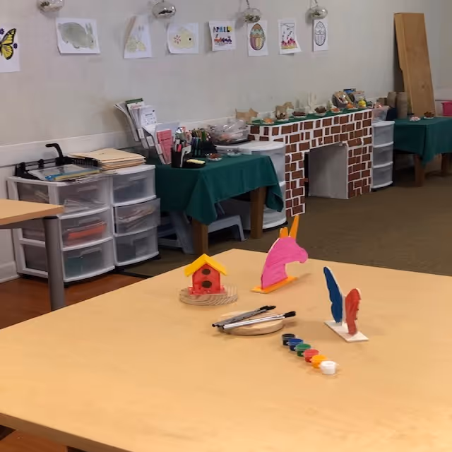A craft room with tables and art supplies including paint, brushes, and small wooden figures. The walls have children's drawings and there are storage drawers and tables covered with green cloths holding various craft materials. A faux brick fireplace decoration is visible in the background.