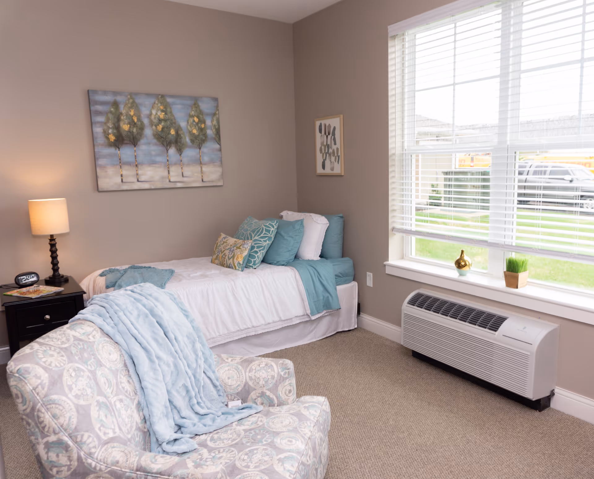 A cozy bedroom with a single bed adorned with white and teal bedding and multiple pillows. A patterned armchair with a light blue throw blanket is positioned in the foreground. A nightstand with a lamp and an alarm clock sits beside the bed. The room has beige walls with two framed artworks and a large window with white blinds letting in natural light. Below the window is a white heating/cooling unit.