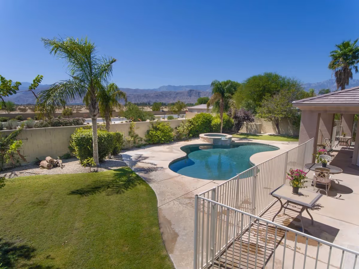 Outdoor view of a backyard area at Desert Hope Elderly care facility featuring a kidney-shaped swimming pool with an attached hot tub, surrounded by a safety fence. There are palm trees, green bushes, and a well-maintained lawn. A covered patio area with tables and chairs is visible on the right side. Mountains and clear blue sky are in the background.