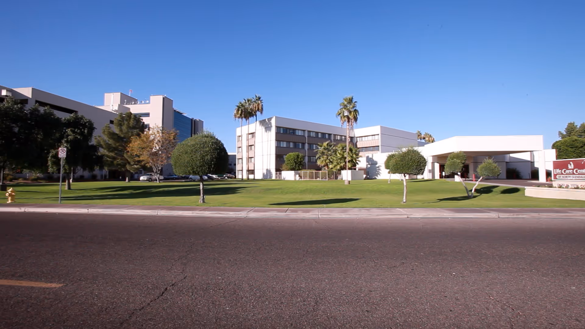 Exterior view of Life Care Center of North Glendale, showing a multi-story building with palm trees and neatly trimmed bushes on a green lawn under a clear blue sky.