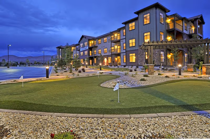Three-story residential building at dusk with lit windows and a landscaped yard featuring a putting green in the foreground.