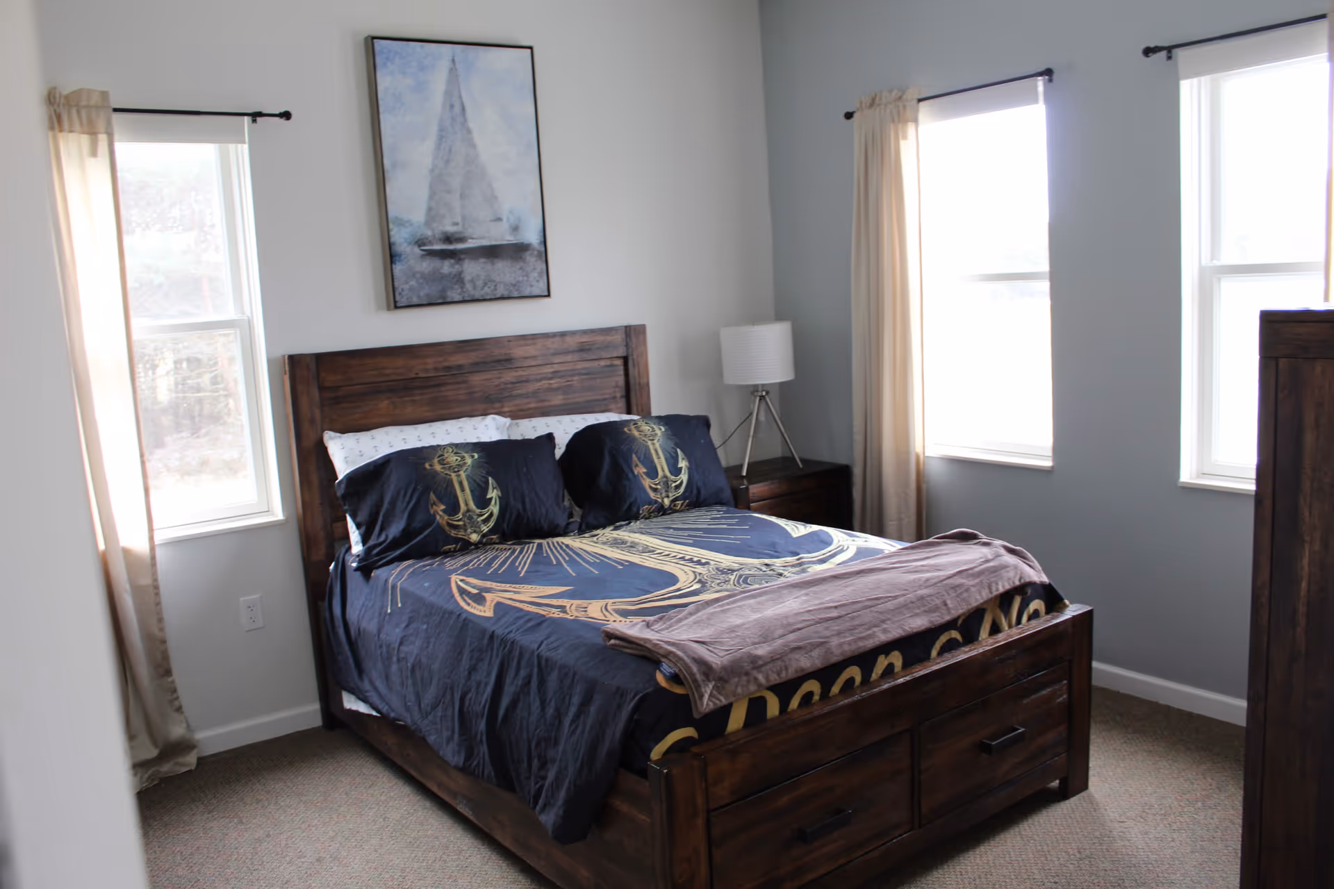 A bedroom with a wooden bed frame and matching nightstand. The bed is made with dark blue bedding featuring anchor designs and a folded brown blanket at the foot. There are three windows with beige curtains letting in natural light. A framed picture of a sailboat hangs on the wall above the bed, and a white lamp sits on the nightstand.