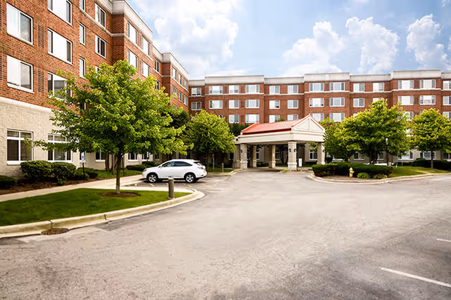 Exterior view of a multi-story senior living facility with a covered entrance, surrounded by green trees and a parking area with a white car parked near the entrance. The building has many windows and a brick and stone facade under a partly cloudy sky.