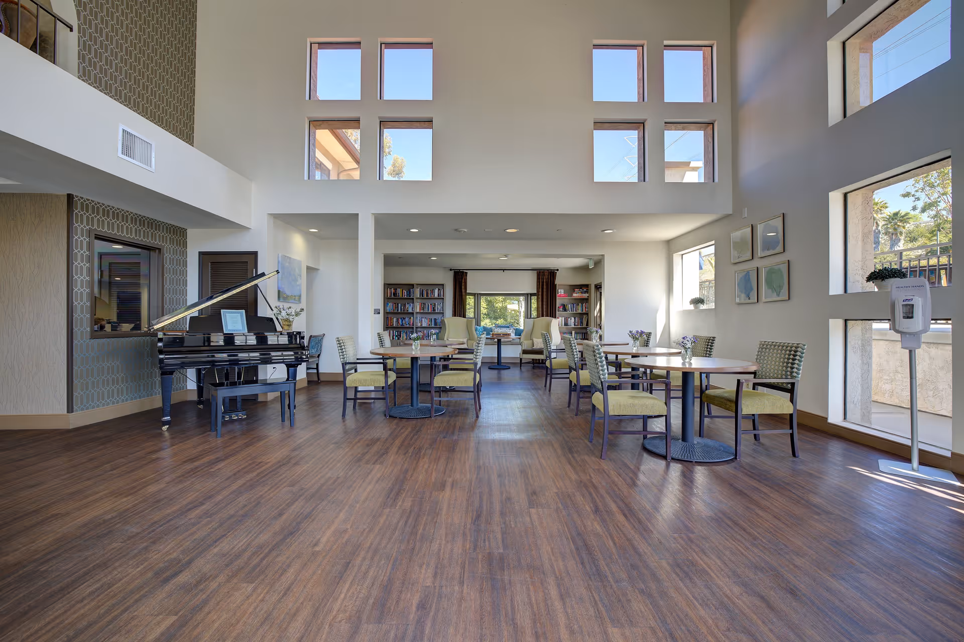 A spacious senior living common area with high ceilings and multiple square windows letting in natural light. The room features several round tables with green cushioned chairs, a black grand piano on the left side, bookshelves in the back, and a hand sanitizer dispenser near the windows on the right.