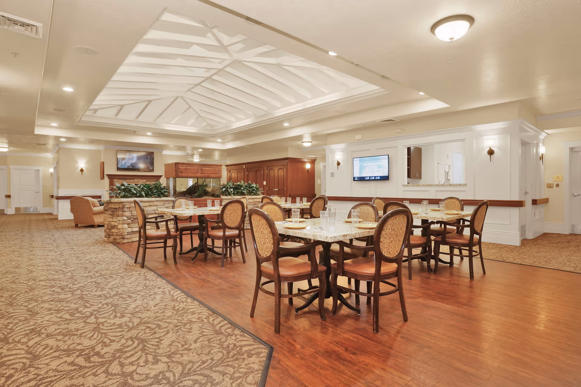 A spacious dining area in a senior living facility with several tables and chairs arranged neatly. The room features a patterned carpet and wooden flooring, a decorative stone planter with greenery, a large ceiling light fixture, and a wall-mounted television. The walls are painted in light colors with wooden accents and sconces providing additional lighting.