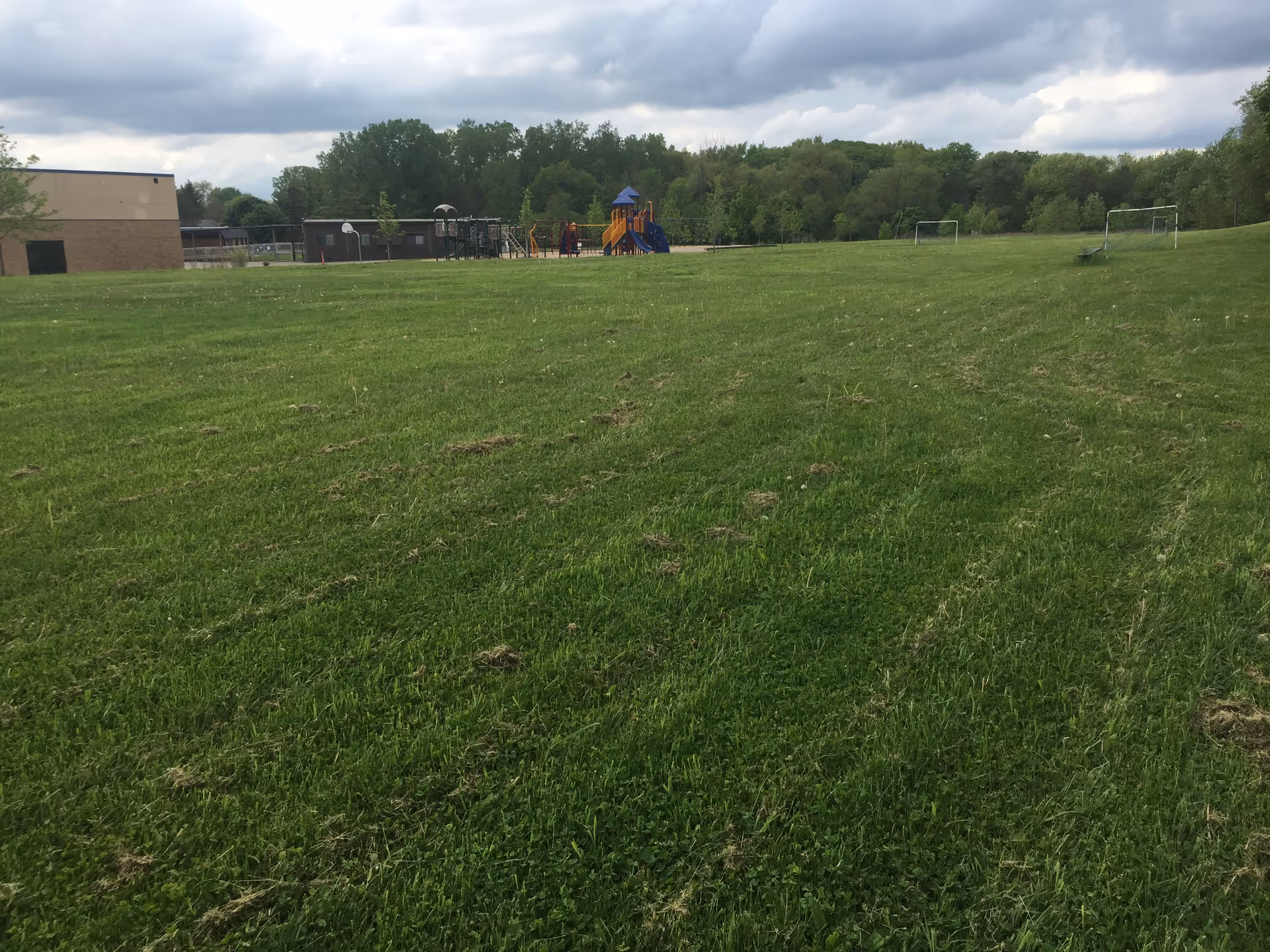 A large grassy field with a playground featuring slides and climbing structures in the distance. There are two soccer goals on the right side of the field and a building on the left. Trees and a cloudy sky are visible in the background.