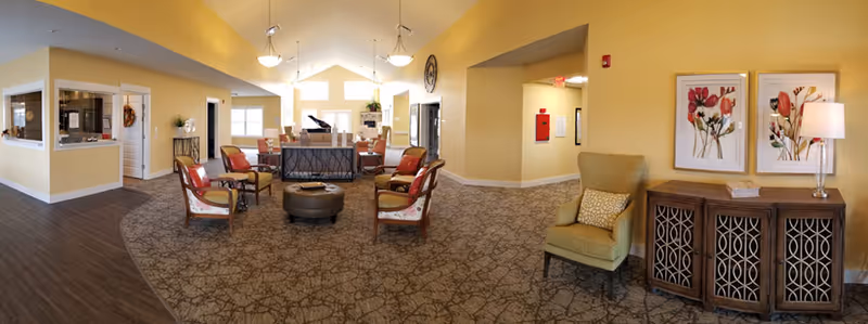 A spacious and well-lit living room area in an assisted living community with yellow walls and a patterned carpet. The room features several armchairs arranged around a central ottoman, a piano in the background, framed floral artwork on the walls, a wooden cabinet with a lamp, and large windows letting in natural light.