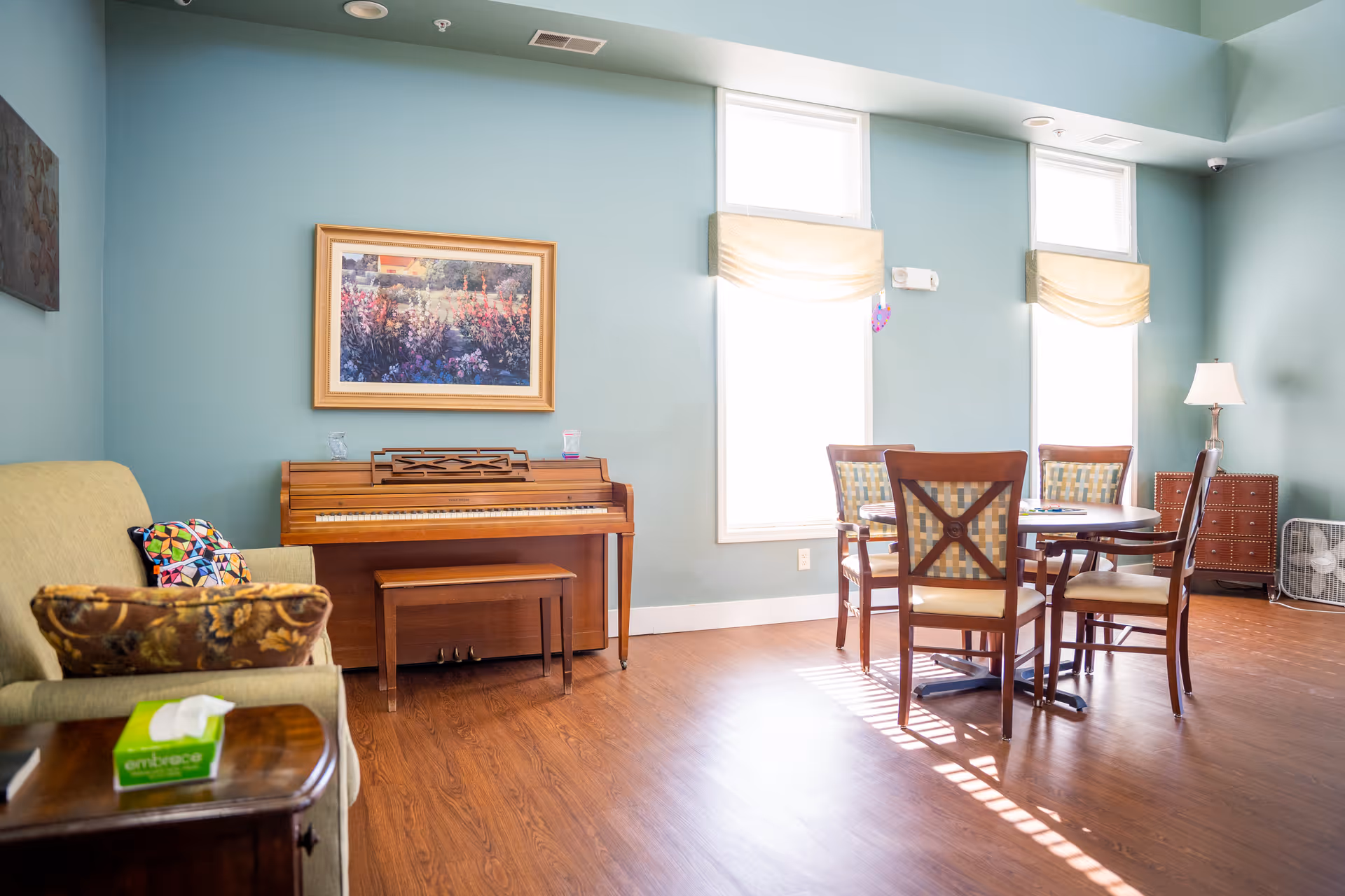 A bright room with light blue walls featuring a wooden piano with a bench, a framed floral painting above it, a round wooden table with four chairs, a beige couch with colorful pillows, a wooden side table with a box of tissues, a floor lamp, and two tall windows with cream-colored shades letting in sunlight.