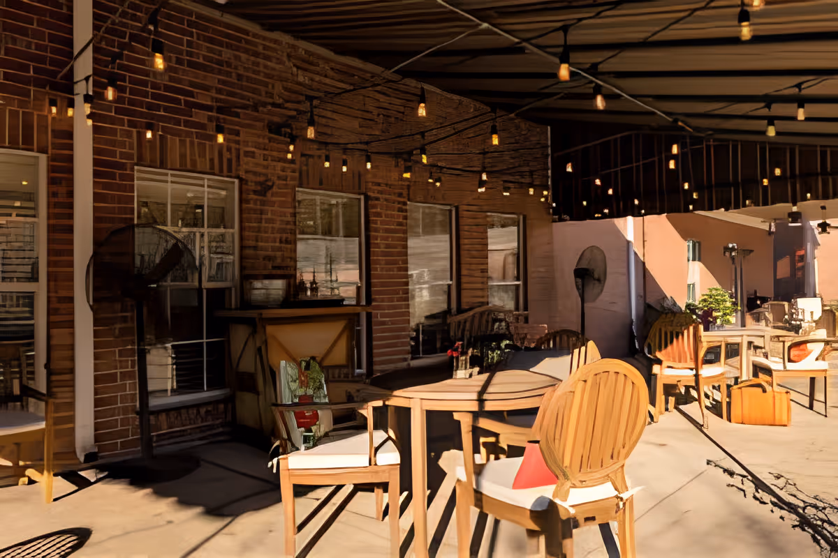 Shaded outdoor patio with wooden tables and chairs, string lights overhead, and a brick building wall with windows.
