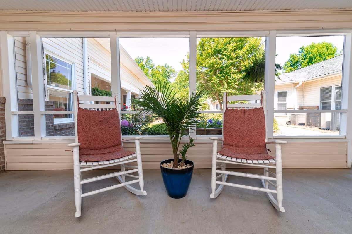 Two white wooden rocking chairs with red cushions placed on a covered porch with large windows overlooking a garden area. A potted green plant in a blue pot is positioned between the chairs.