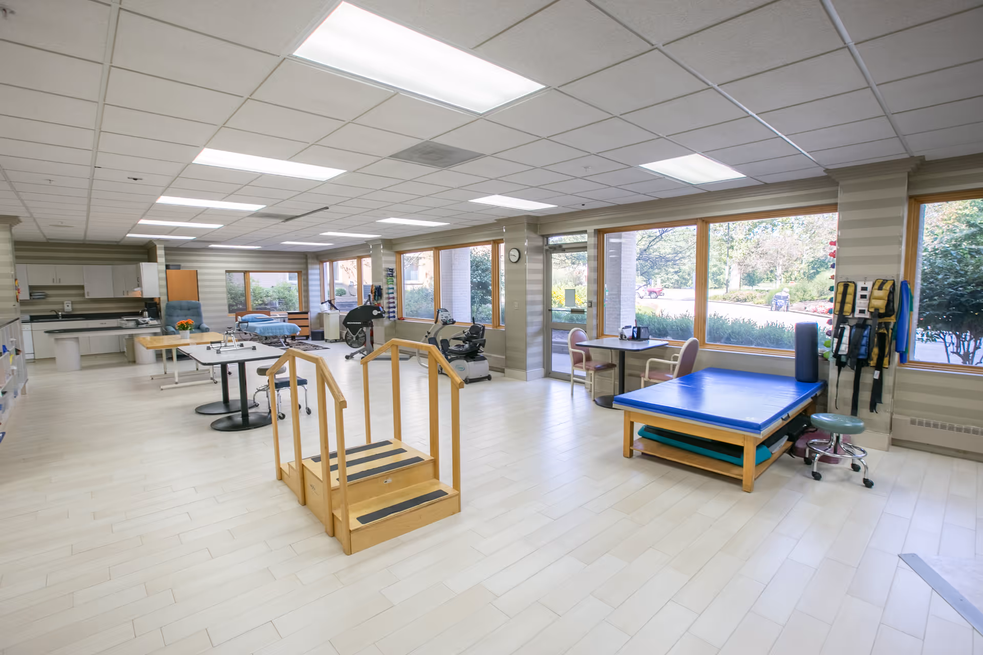 A spacious physical therapy room with large windows letting in natural light. The room contains exercise equipment including stationary bikes, a set of wooden stairs for rehabilitation, a padded therapy table, and various therapy tools mounted on the walls. There is also a small table with chairs near the windows and a kitchenette area in the back.