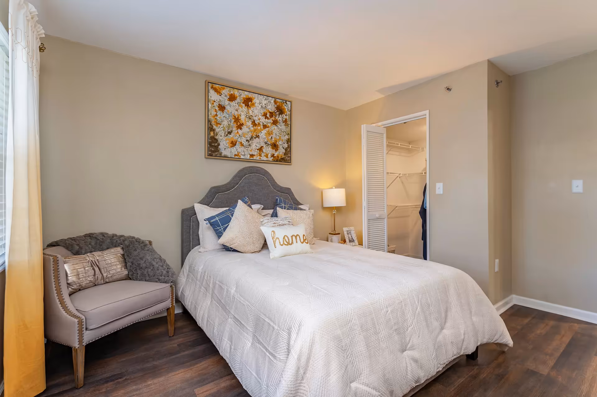 A cozy bedroom with a neatly made bed featuring multiple pillows, including one with the word 'home' embroidered on it. There is a cushioned armchair with a throw blanket and pillow next to a window with sheer curtains. A bedside table with a lamp and framed photos is beside the bed. The room has wooden flooring, beige walls, and a floral painting above the bed. An open door reveals a walk-in closet with shelving and hanging space.