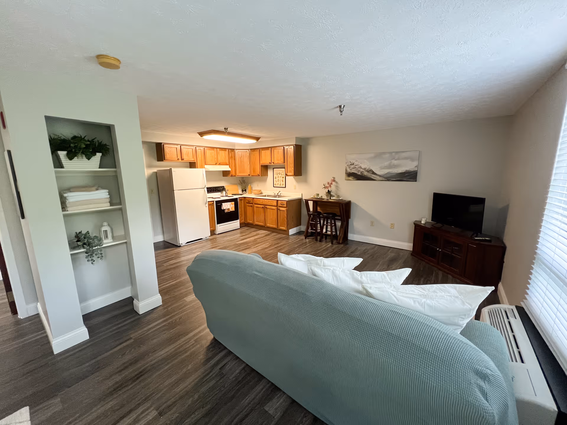 A cozy senior living community interior featuring a small kitchen with wooden cabinets, a white refrigerator, stove, and sink. In front of the kitchen is a light blue couch with white pillows facing a TV on a wooden stand. There is a small dining table with two chairs against the wall, a window with blinds, and a built-in shelf with plants and towels.
