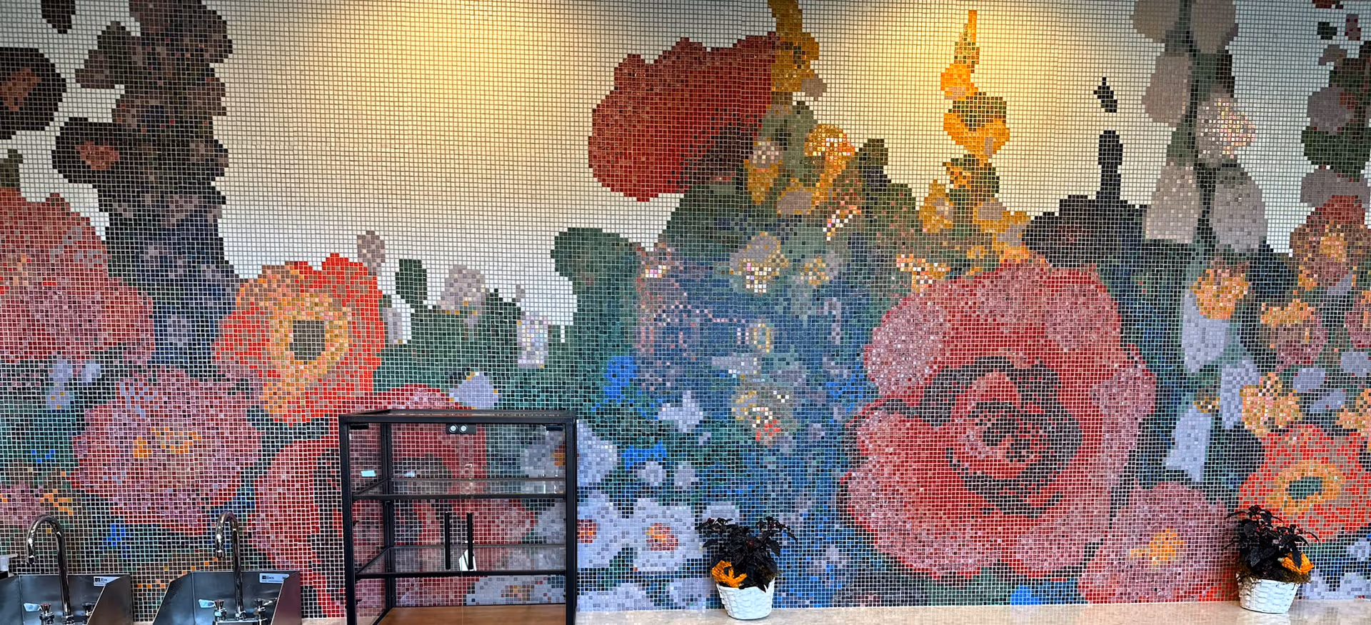 A colorful mosaic wall featuring large flowers in shades of red, pink, orange, and green. In front of the wall, there are two sinks with faucets on the left side, a black metal shelving unit in the center, and two small potted plants with orange flowers on the right side.
