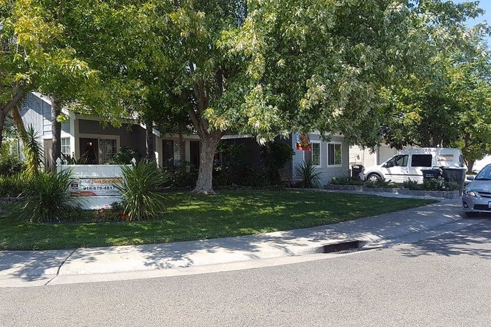 Street-front view of a single-story elder care home with large trees, a green lawn, a front sign, and parked vehicles.