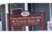 Wooden sign reading "Fiddlers Green Manor Nursing Home - Pride in Caring" mounted in front of a gray building.