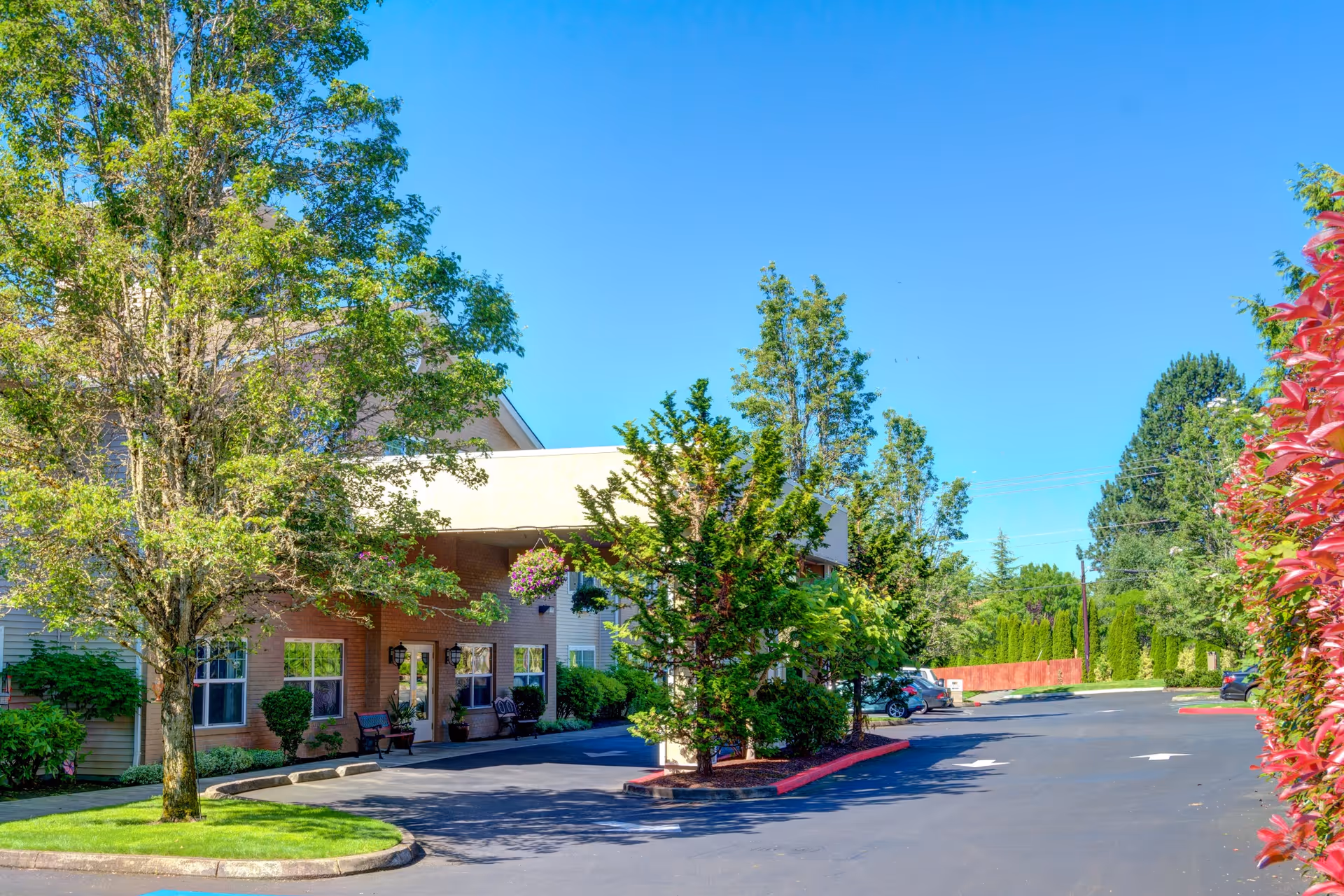 Exterior view of Princeton Village by Cogir showing the entrance area with trees, shrubs, and a parking lot under a clear blue sky.