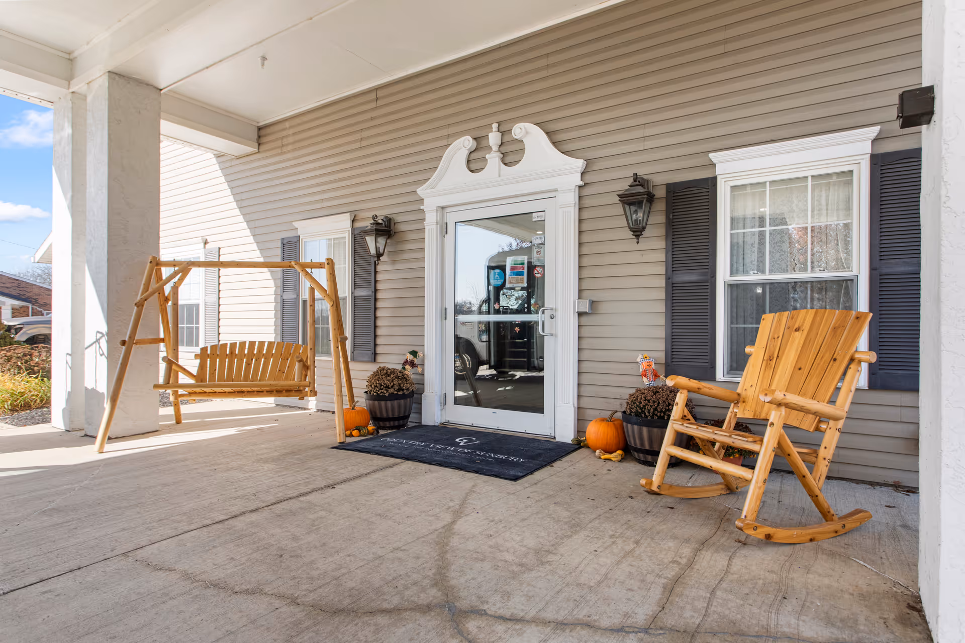 Covered porch area at the entrance of a building with a wooden swing bench on the left and a wooden rocking chair on the right. The entrance door is glass with white trim and flanked by two windows with black shutters. There are two potted plants with pumpkins placed near the door, and two wall-mounted lantern-style lights on either side of the door.