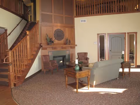 A cozy senior living facility common area with a wooden staircase on the left, a fireplace with decorative items above it, two upholstered armchairs, a sofa, and wooden side tables with candles. The room has beige walls and carpeted flooring with a patterned design.