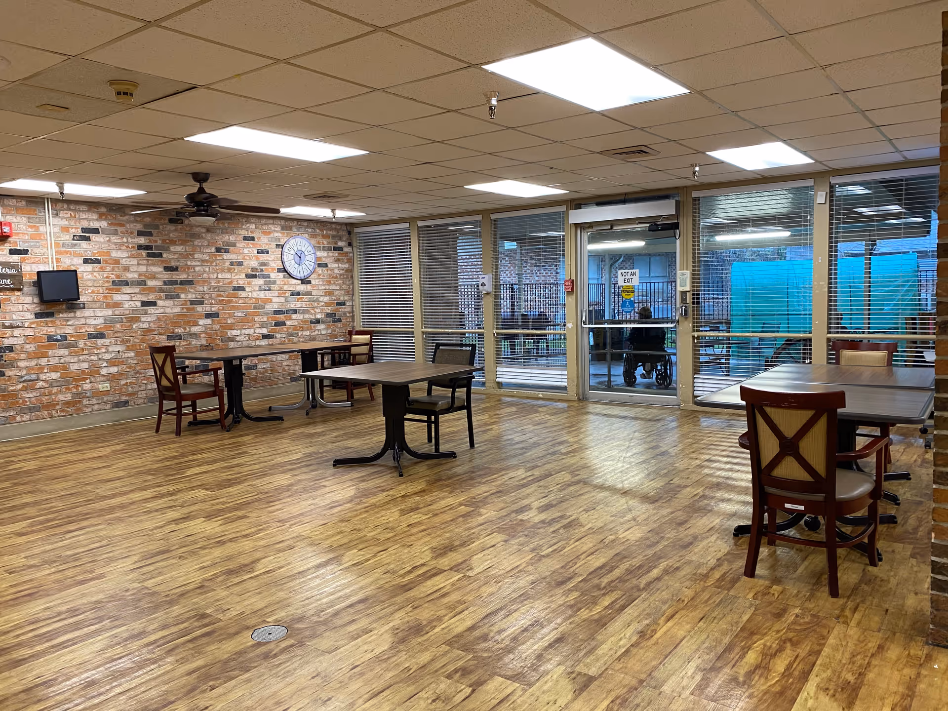 A spacious room with wooden flooring and brick walls, furnished with several tables and chairs. Large windows and glass doors with blinds allow natural light to enter, and a clock is mounted on the brick wall. The room appears to be a common area or dining space in a senior living facility.