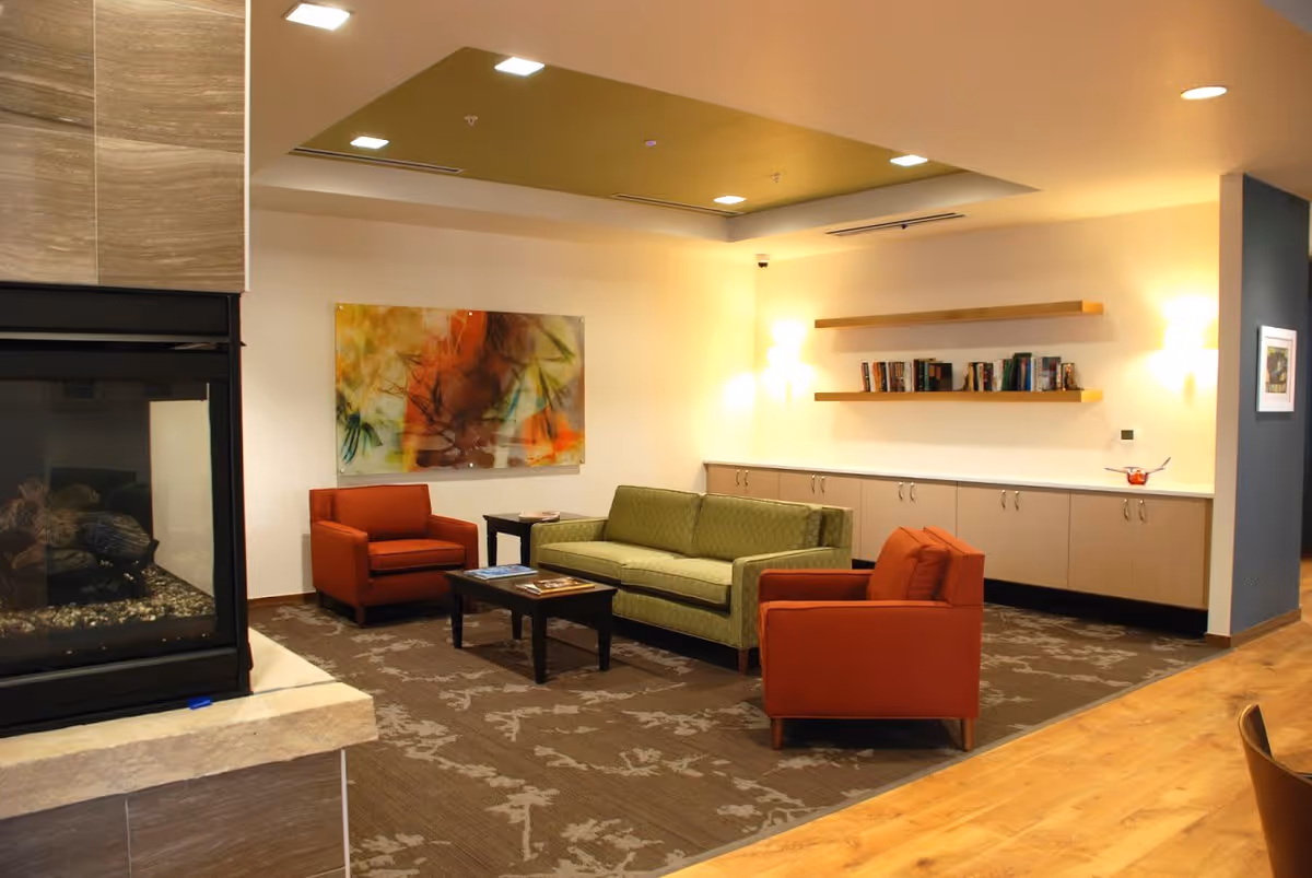 A cozy living room area with a green sofa and two orange armchairs arranged around a dark wooden coffee table. There is a modern fireplace on the left side and a wall with two wooden shelves holding books and a long cabinet below. The room has warm lighting and a decorative abstract painting on the wall.