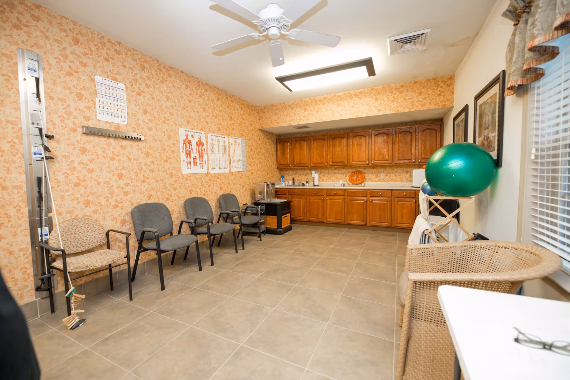A therapy or exercise room with floral wallpaper on one wall and white paint on the others. There are four gray chairs and one patterned chair lined up against the floral wall, which has anatomical charts displayed. The opposite wall has wooden cabinets with a countertop holding various items. A green exercise ball is placed on a wooden rack near a window with blinds and a wicker chair. The floor is tiled, and the ceiling has a white fan and a rectangular light fixture.