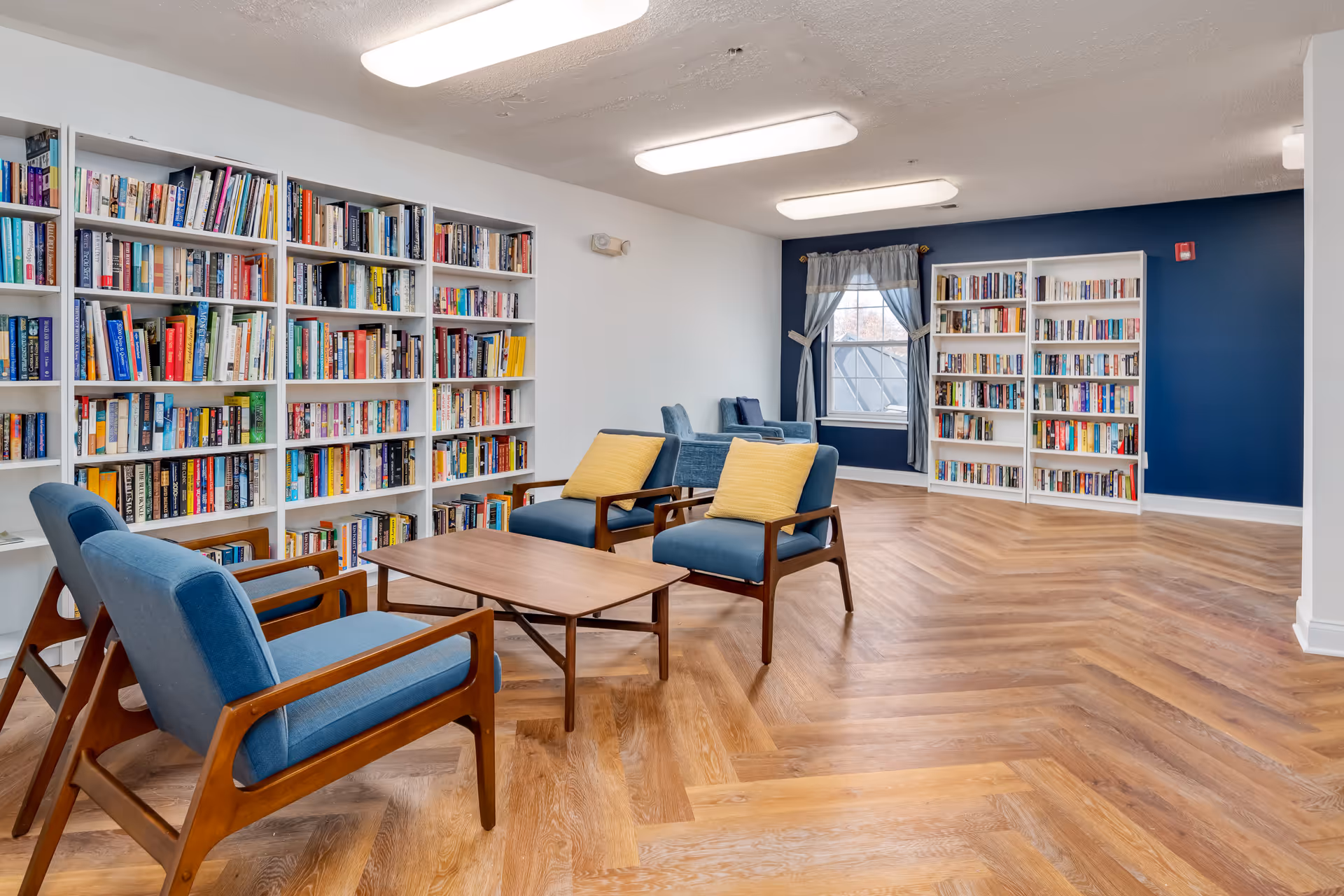 Bright communal reading room with bookshelves, mid-century blue armchairs, a wooden coffee table and herringbone wood flooring.