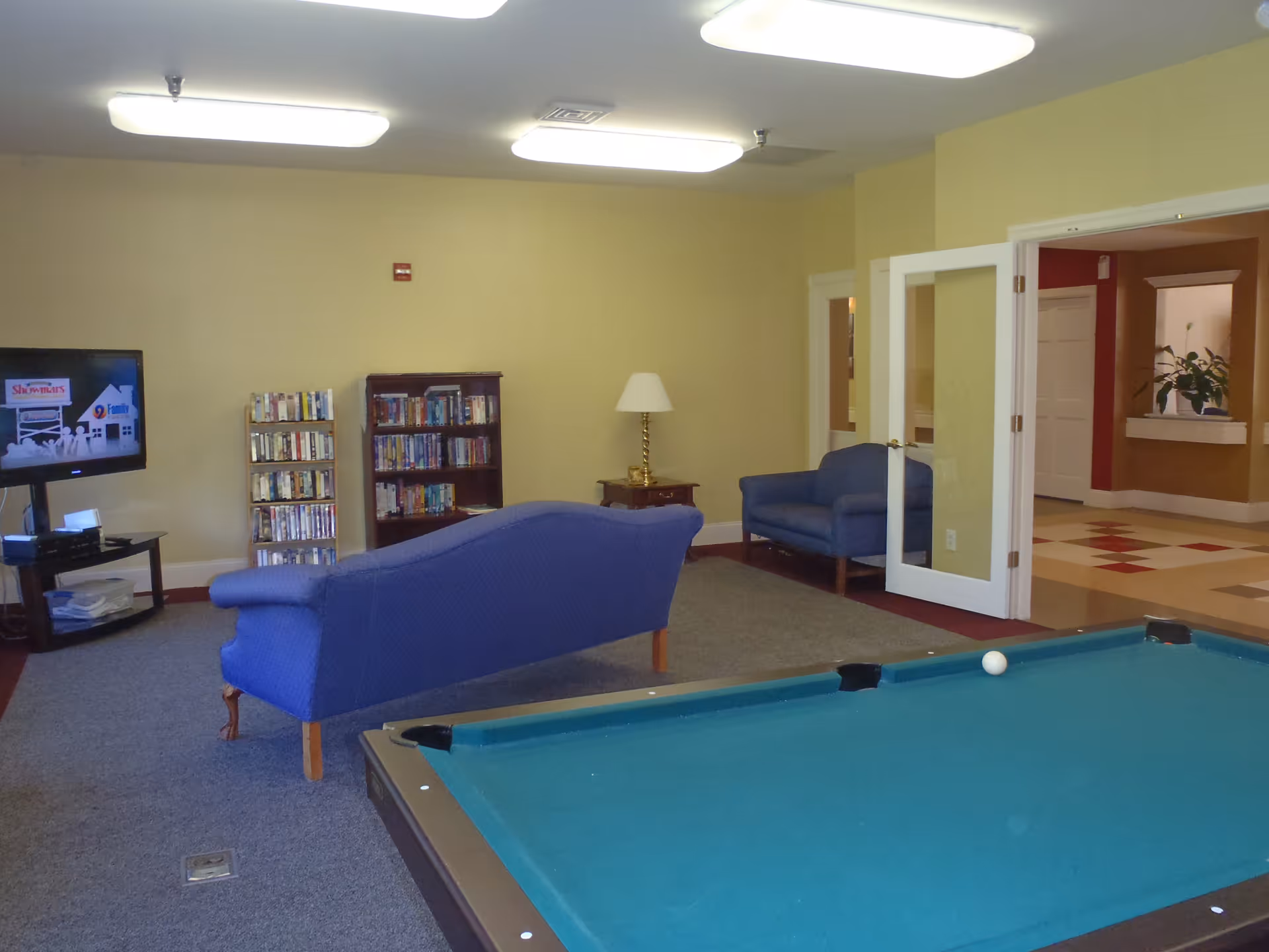 Recreation room with a pool table in the foreground, blue sofas, a TV, and bookshelves.