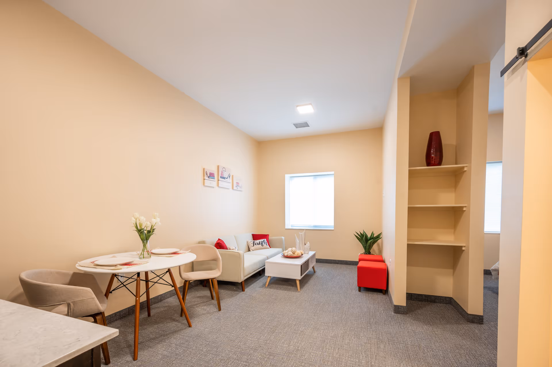 Small furnished living area with a round dining table and chairs, a sofa and coffee table, shelving and neutral walls.