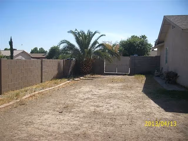 Dirt backyard with a palm tree, cinderblock fence, a gated fence at the far end, and the side of a house under a clear sky.