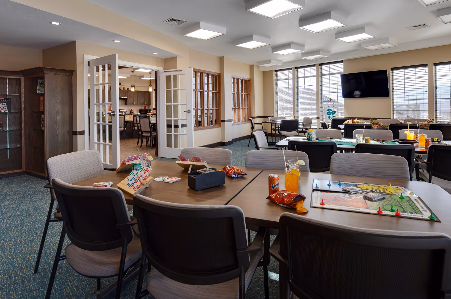 Spacious activity/dining room with tables and chairs set up for games and snacks, large windows and a wall-mounted TV.