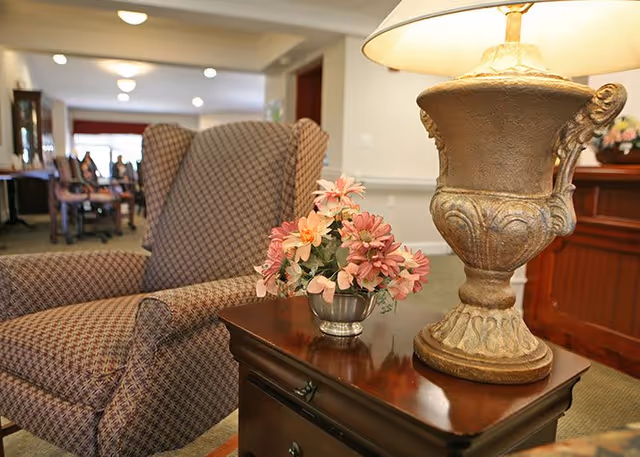 A cozy seating area in a senior living facility featuring a patterned armchair next to a wooden side table with a decorative lamp and a small vase of pink and peach flowers. In the background, more chairs and tables are visible in a softly lit hallway or common area.