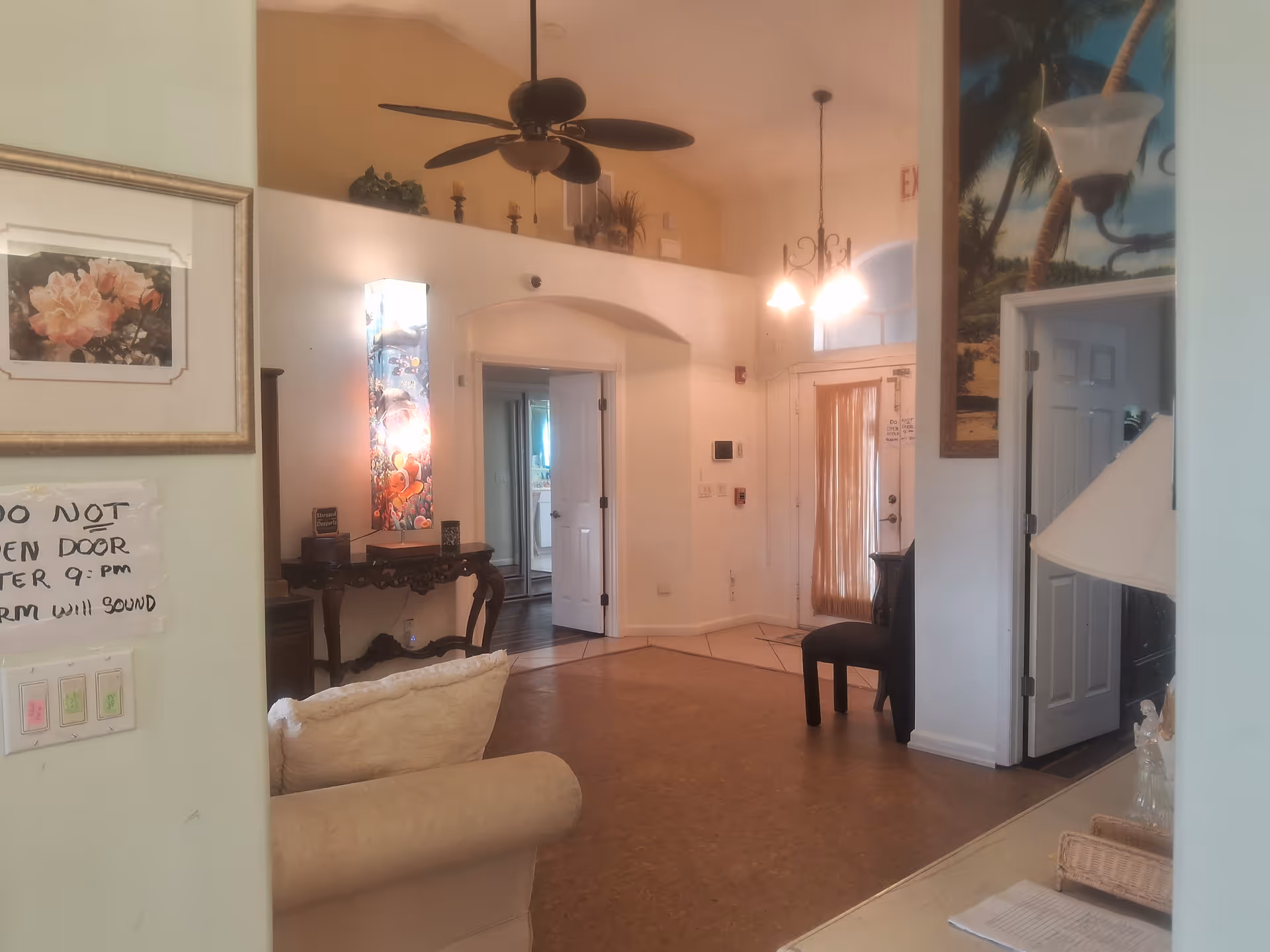 Interior view of a living area in Violet Gardens Assisted Living Facility featuring a beige couch, a decorative table with a colorful lamp, a ceiling fan, a framed floral picture, and a door with a curtain. There is a sign on the wall near light switches that reads 'DO NOT OPEN DOOR AFTER 9:00 PM ALARM WILL SOUND.'