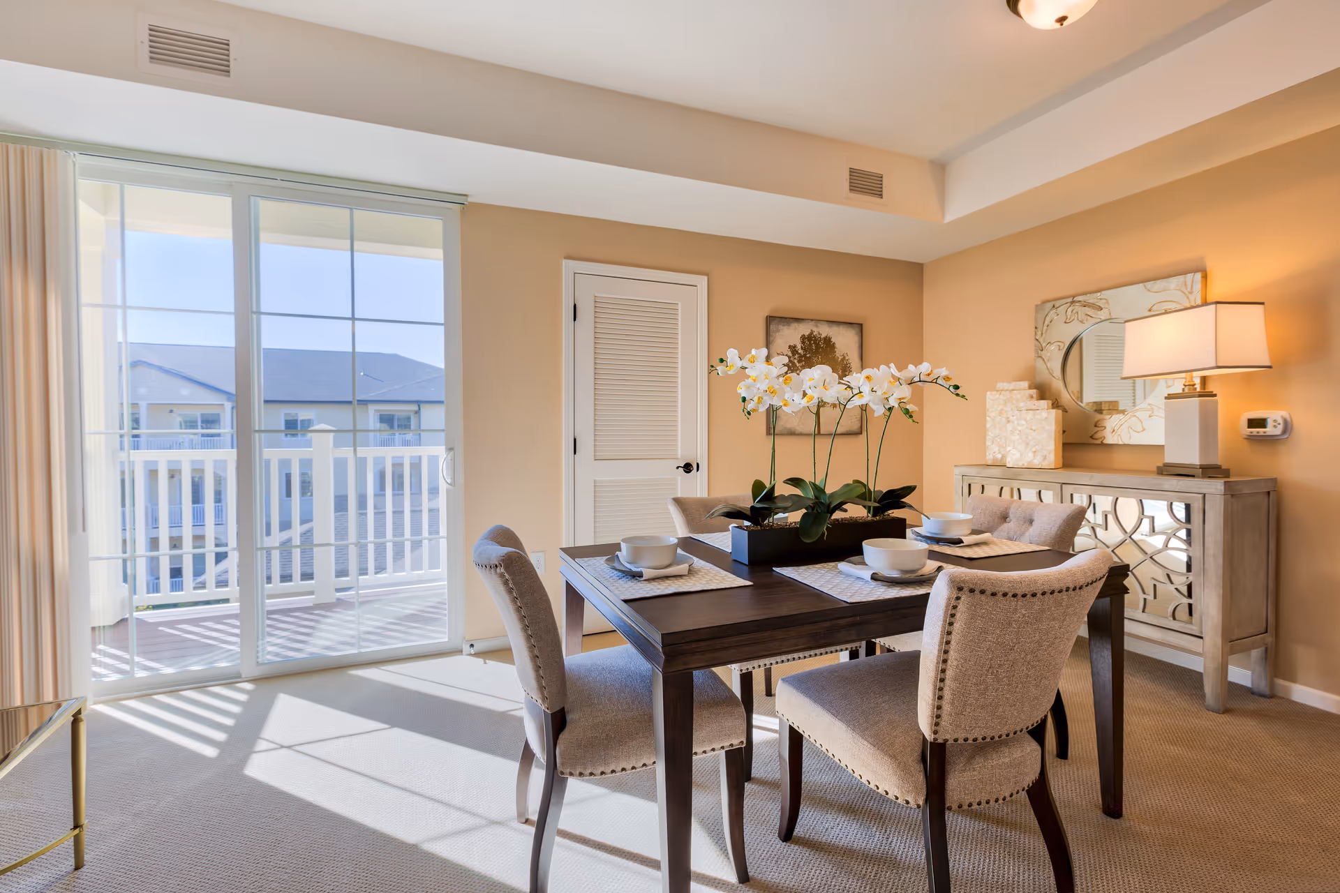 A bright dining room with a dark wooden table set for four with white bowls and napkins. The table has a centerpiece of white orchids. Four upholstered chairs surround the table. There is a sliding glass door leading to a balcony with a white railing. A sideboard with a lamp, decorative items, and a mirror hangs on the wall. The room has beige walls and carpeted floor.