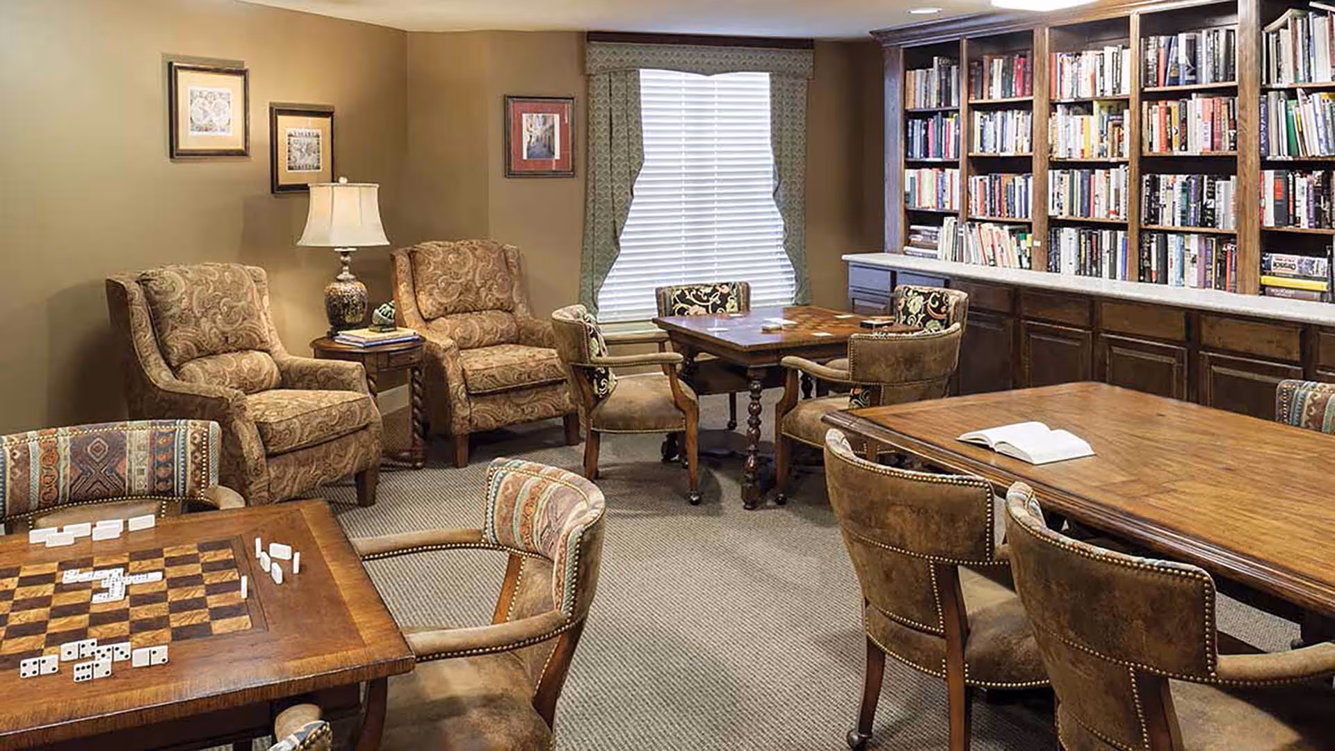 A cozy room with multiple wooden tables and upholstered chairs arranged for games and reading. One table has a checkerboard with dominoes on it. There are two patterned armchairs with a side table and lamp between them. A large bookshelf filled with books lines one wall, and a window with blinds and a decorative valance is in the background.