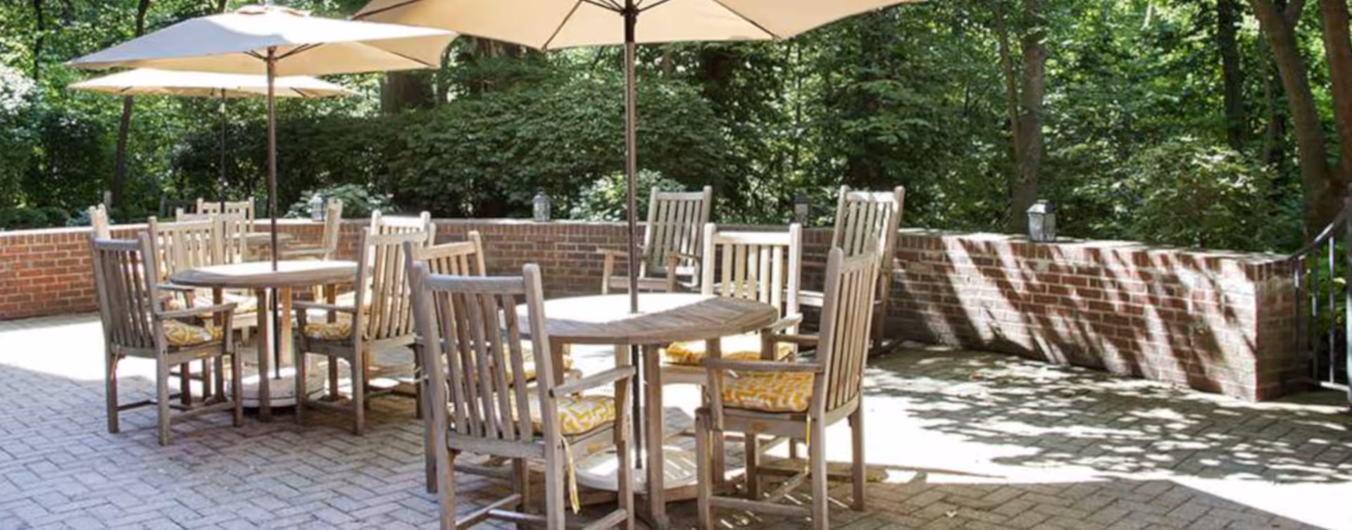 Outdoor patio with wooden tables and chairs under umbrellas beside a low brick wall and trees.