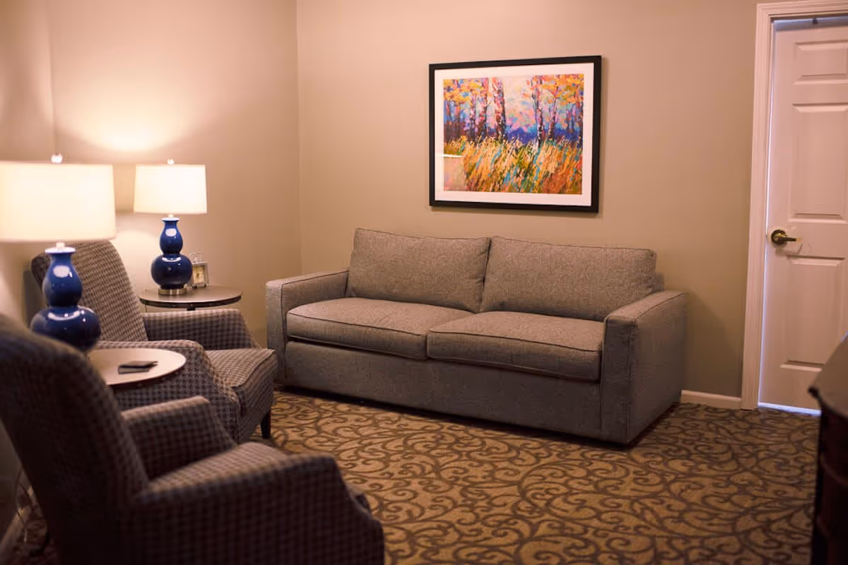 A cozy living room with a gray sofa, two patterned armchairs, two blue table lamps on round side tables, a colorful framed painting on the beige wall, and a patterned carpet. A closed white door is visible on the right side.