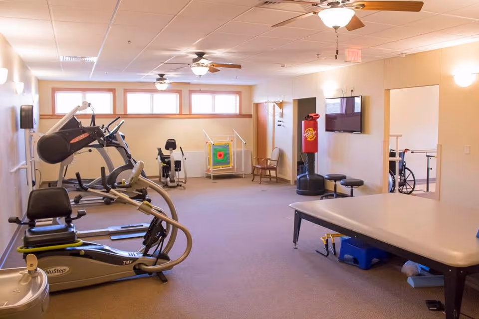 A spacious exercise room in an assisted living facility with various fitness equipment including a NuStep machine, an elliptical, a recumbent bike, a punching bag, and a large therapy table. The room has beige walls, carpeted floor, ceiling fans with lights, and windows near the ceiling allowing natural light. There is a wheelchair visible in an adjacent room through an open doorway.