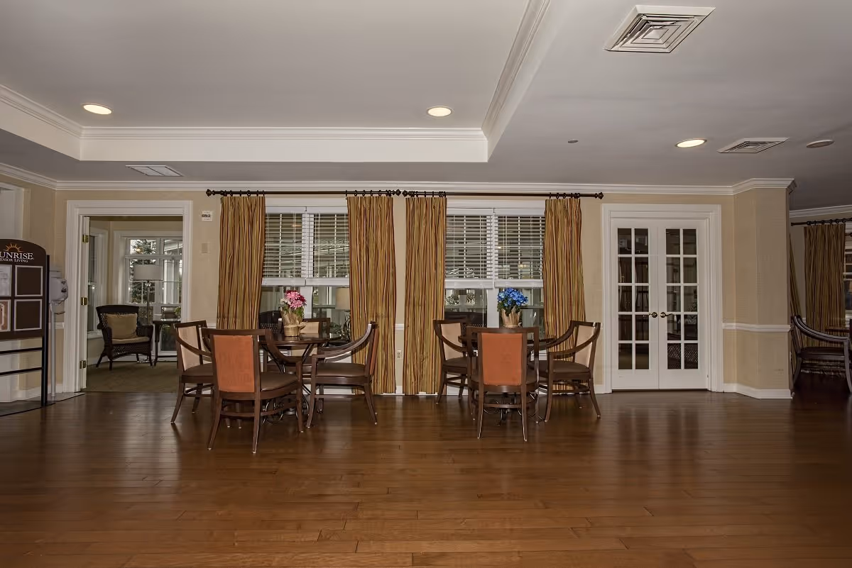 Spacious communal dining room with round tables and chairs on hardwood floors, windows with curtains, and double French doors.