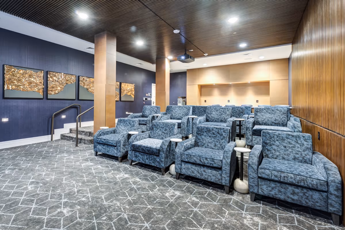 A modern theater room with three rows of blue upholstered armchairs, each with a small round side table. The room has a patterned gray carpet, dark blue walls with gold abstract artwork, wooden paneling on one side, and a dark wood slatted ceiling with recessed lighting. There is a projector mounted on the ceiling and steps leading up to the seating area.